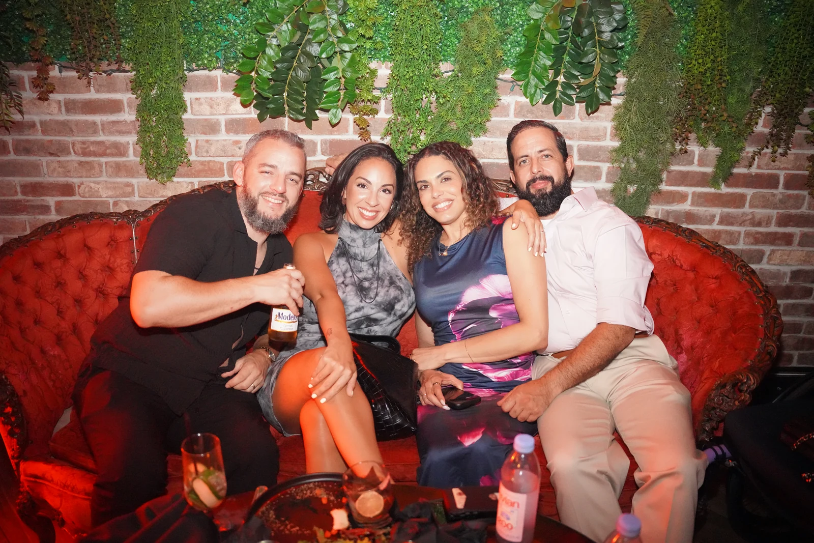 Four smiling guests seated together on a red banquette beneath hanging greenery
